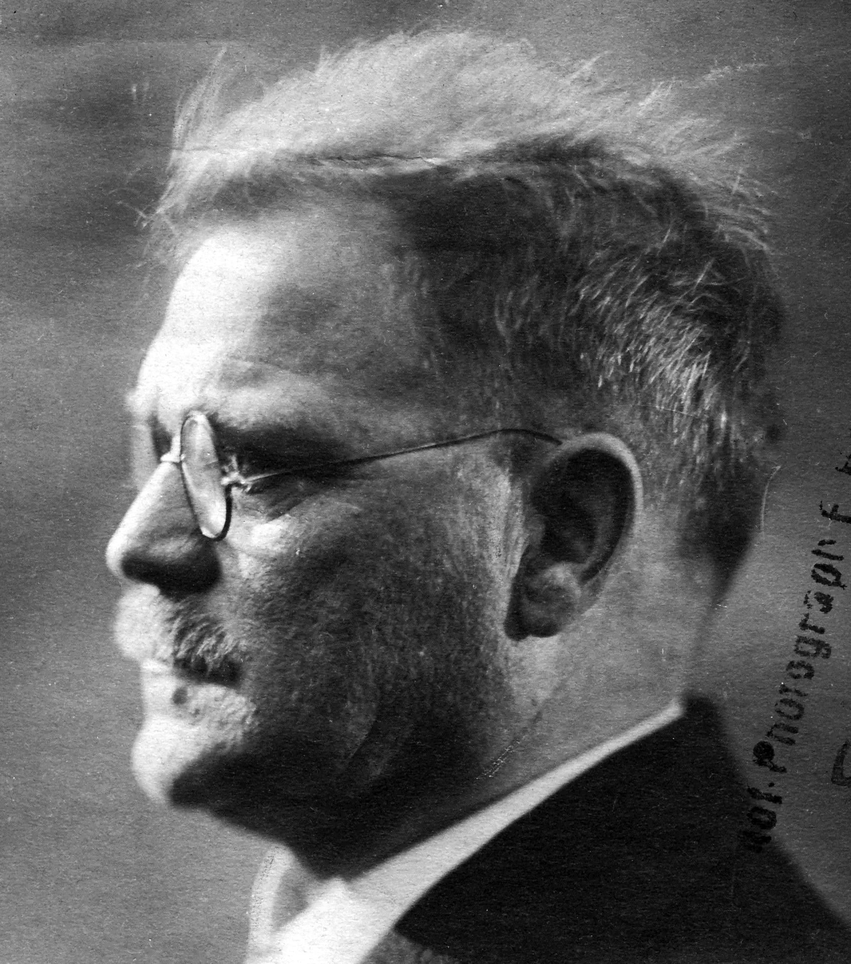 A black and white profile view of a man with a moustache, spectacles, and dishevelled hair.