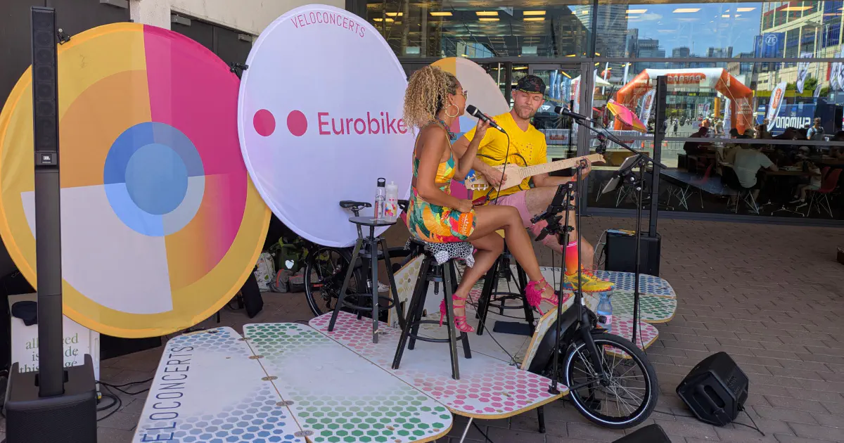 A music stage with a singer and a guitar player. The stage is foldable and mounted to a purpose built carbo bike. The Eurobike banner is in the background.