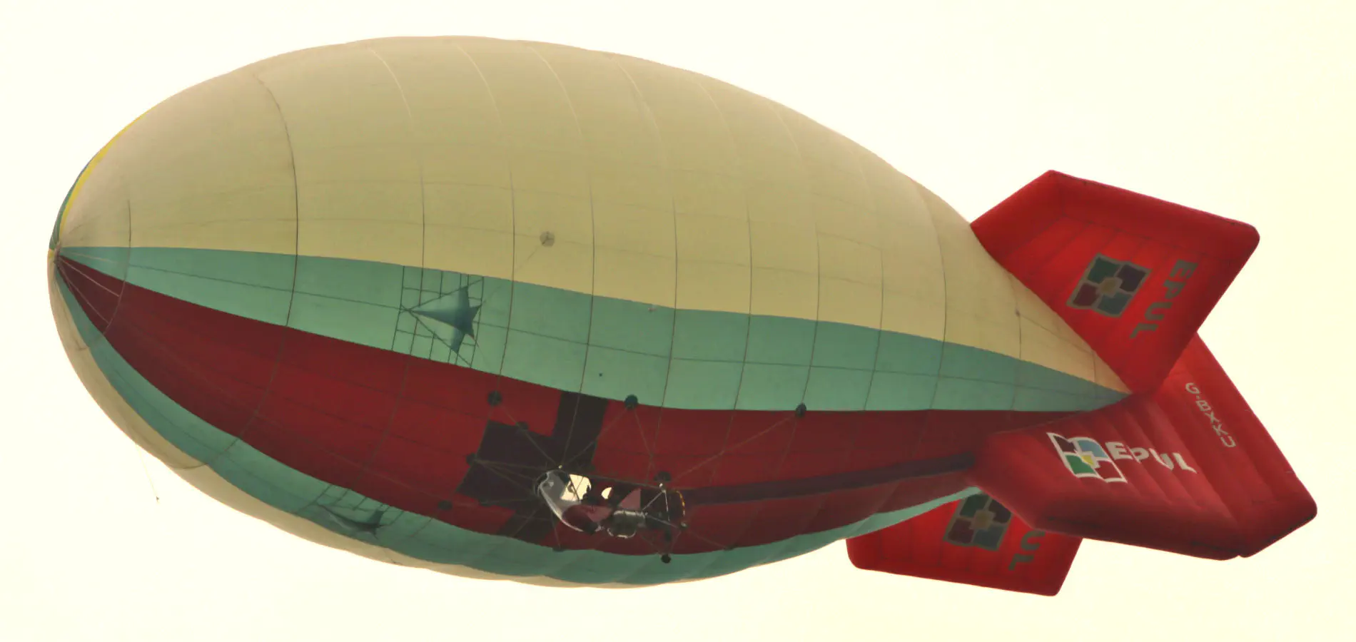 The thermal airship from underneath. The gondola, which seats two persons, has the engine mounted at the back (the propeller cage features red and yellow stripes, just about visible in the image). Above the pilot there is a visible cut-out in the hull, with two burners mounted just above it (not visible in the image). The thermal airship’s rudder is deflected to the left for a left turn.
