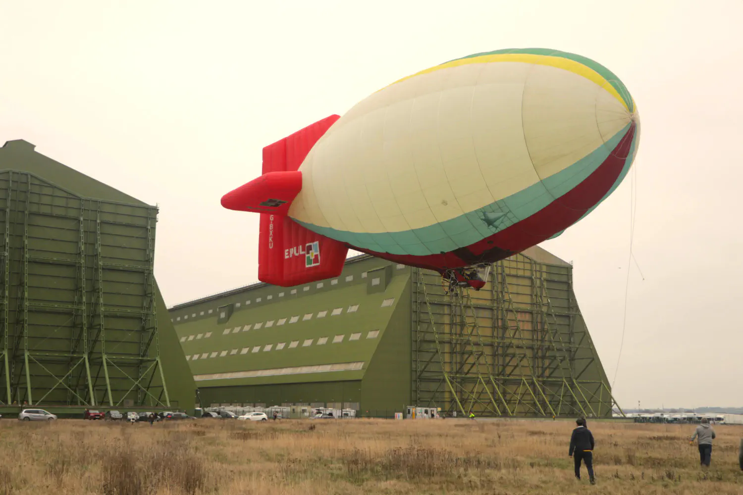 A thermal airship with red tailfins and a mostly white hull with red, blue, yellow, and green elements is floating around 10 m above the ground. In the background, there are two very large green airship sheds.