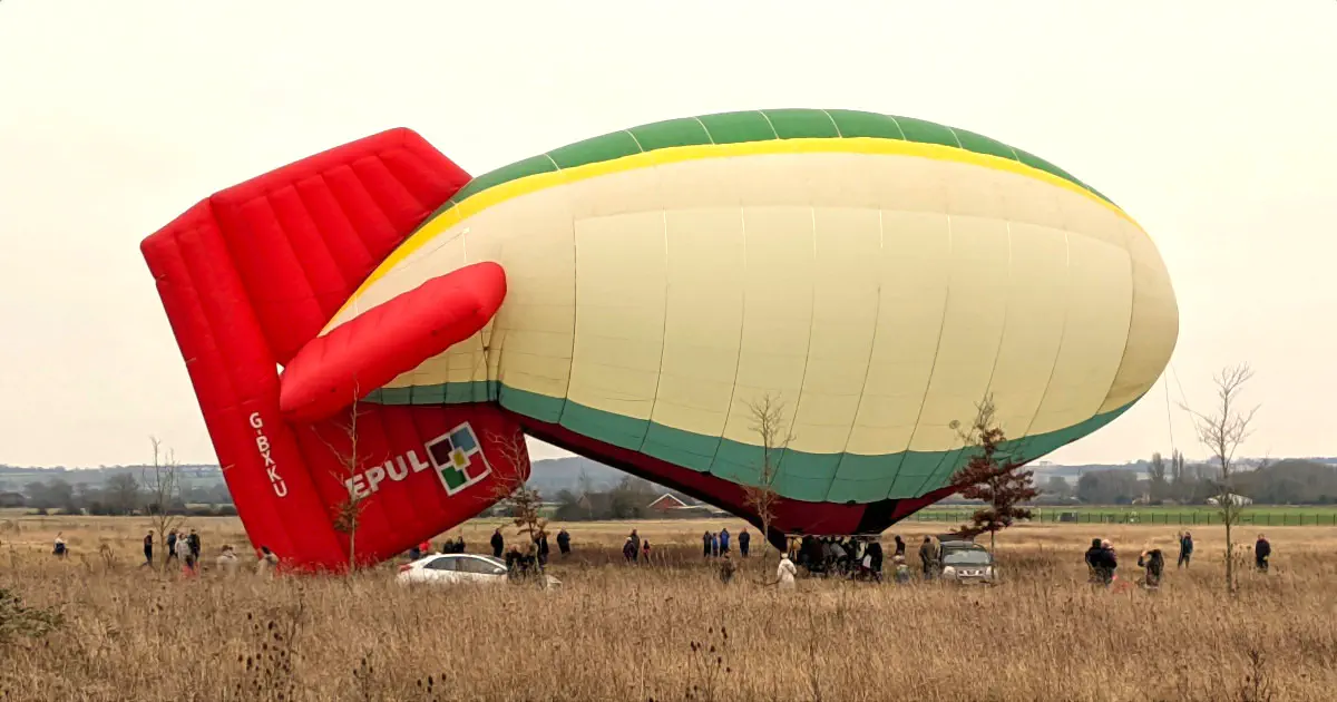 A thermal airship on the ground. Its hull is insufficiently inflated, resulting in the tail drooping. It has red vertical and horizontal stabilisers.