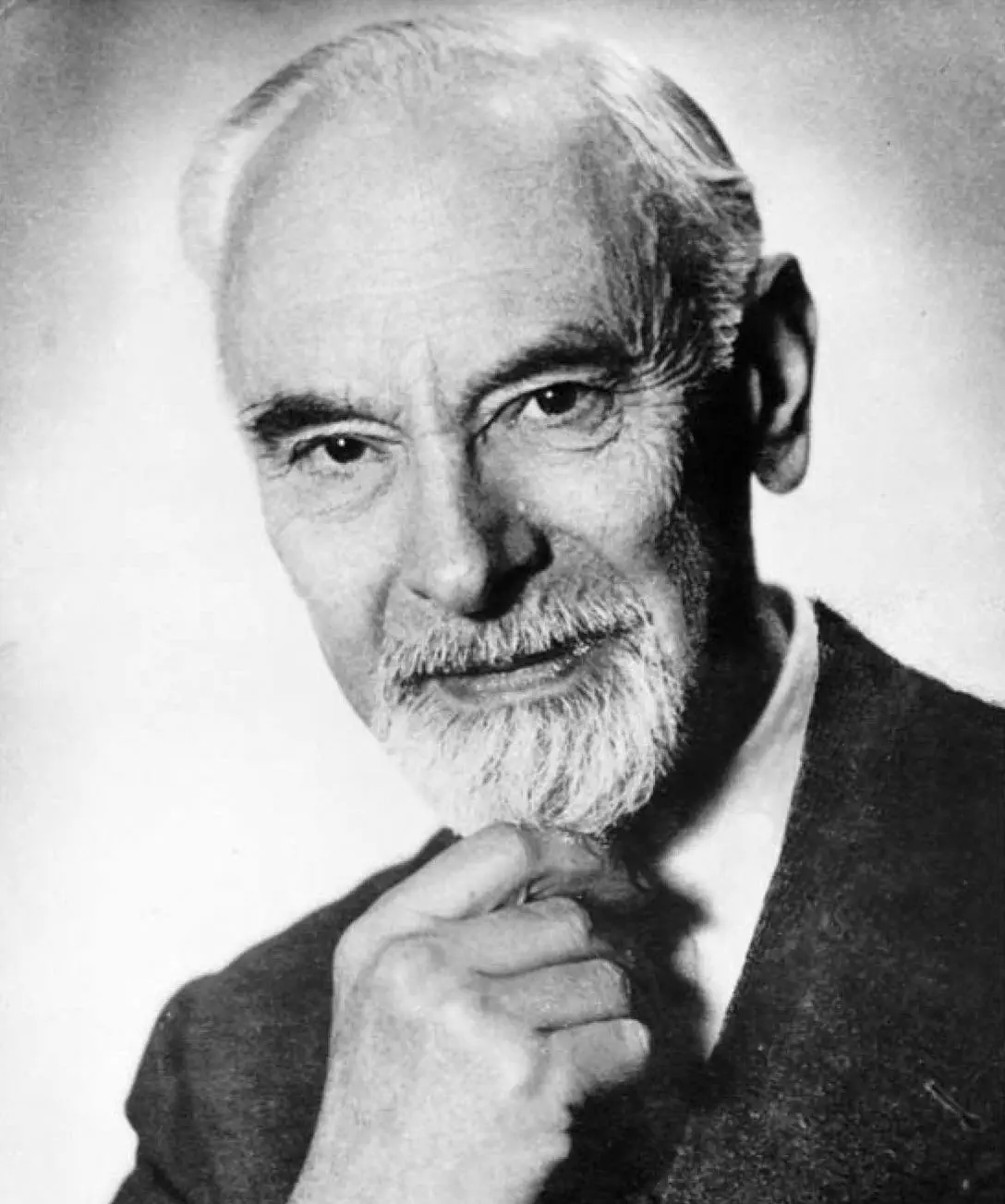 A black and white image of an old man with white hair, an expansive forehead, dark prominent eyebrows, and a short white beard, holding his chin with his right hand. He appears deep in thought and smiles faintly.